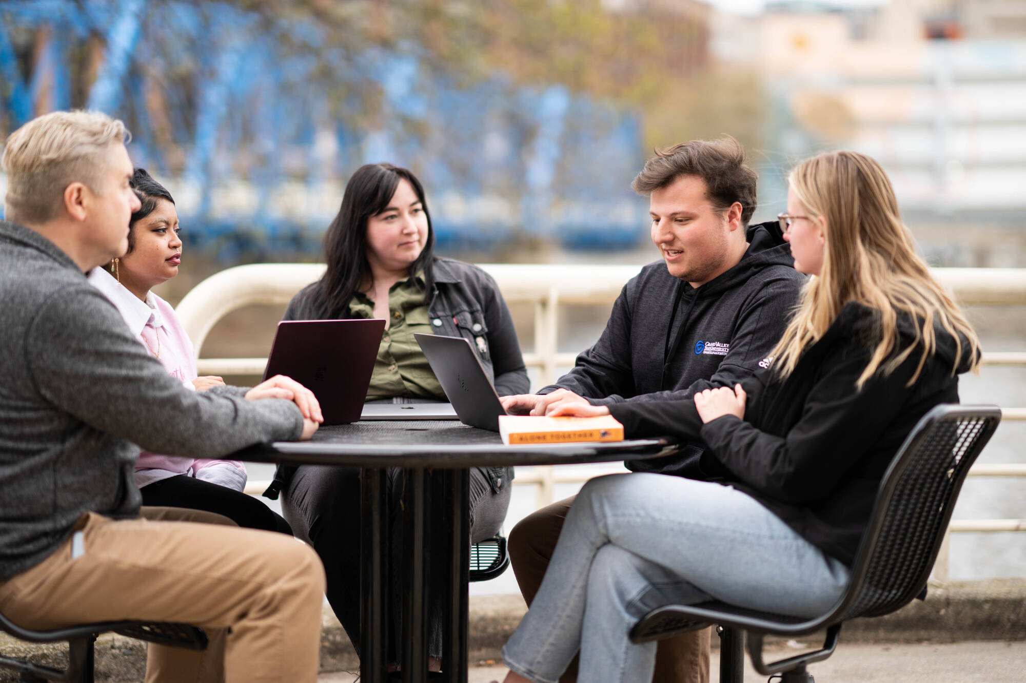 A group of five students sit outdoors at a round table, engaged in discussion with laptops and notebooks open. They appear to be collaborating or studying together, with a blurred cityscape and river in the background.